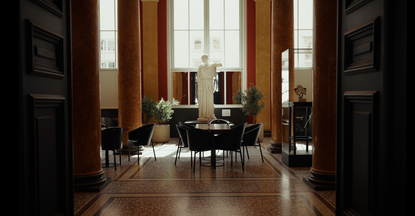 Blick in eleganten Säulenraum mit hohen Decken, Tischen, Stühlen und einer Skulptur