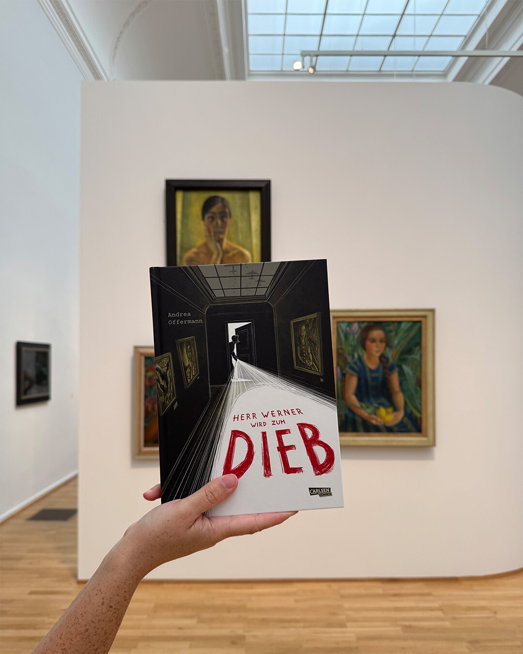 Hand hält Buch mit dem Titel "Herr Werner wird zum Dieb" vor Gemälden in einer Galerie