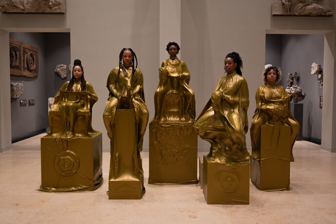 Sechs Frauen in goldenen Kleidern posieren auf großen würfelförmigen Objekten in einer Kunstgalerie