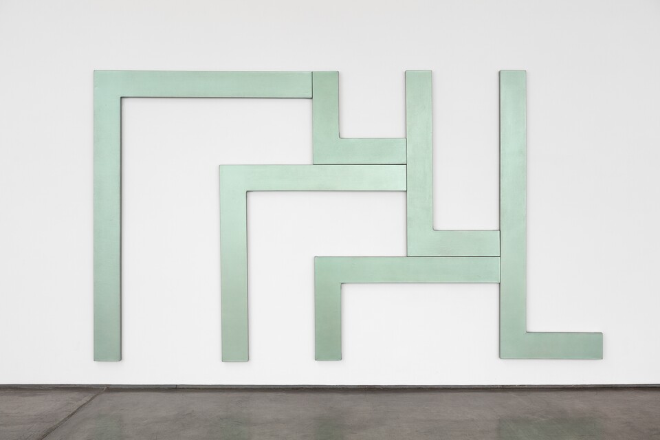 Grüne geometrische Installation an weißer Galeriewand über dunklem Boden
