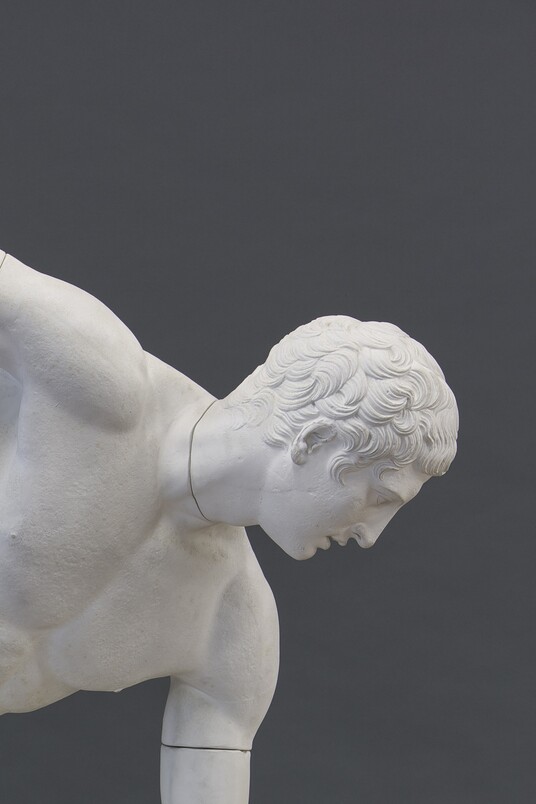 Weißer lebensgroßer Gipsabguss einer Marmorskulptur des Diskuswerfers in klassischer Wurfhaltung vor grauem Hintergrund
