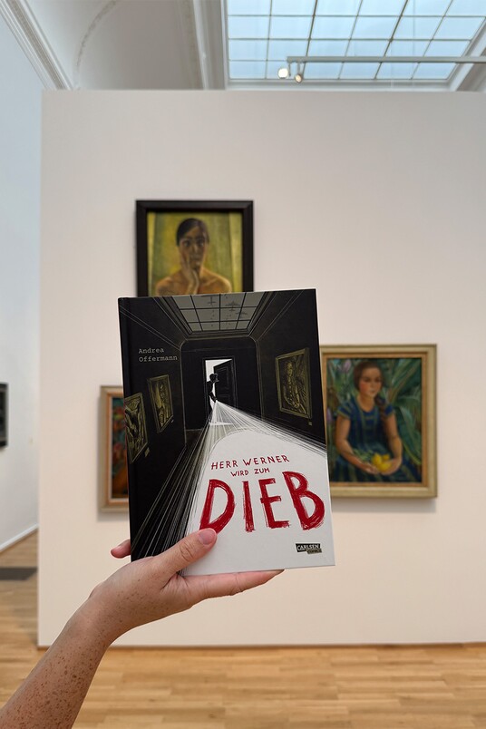 Hand hält Buch mit dem Titel "Herr Werner wird zum Dieb" vor Gemälden in einer Galerie