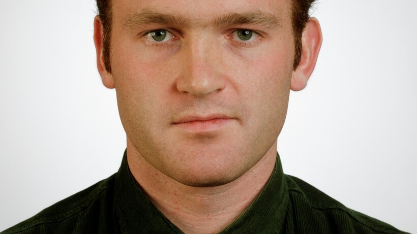 Portraitfoto eines jungen Mannes mit braunem Haar in schwarzem Poloshirt vor weißem Hintergrund