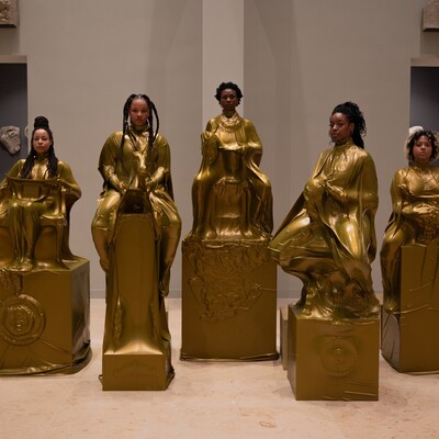 Sechs Frauen in goldenen Kleidern posieren auf großen würfelförmigen Objekten in einer Kunstgalerie