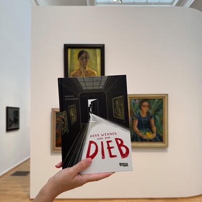 Hand hält Buch mit dem Titel "Herr Werner wird zum Dieb" vor Gemälden in einer Galerie